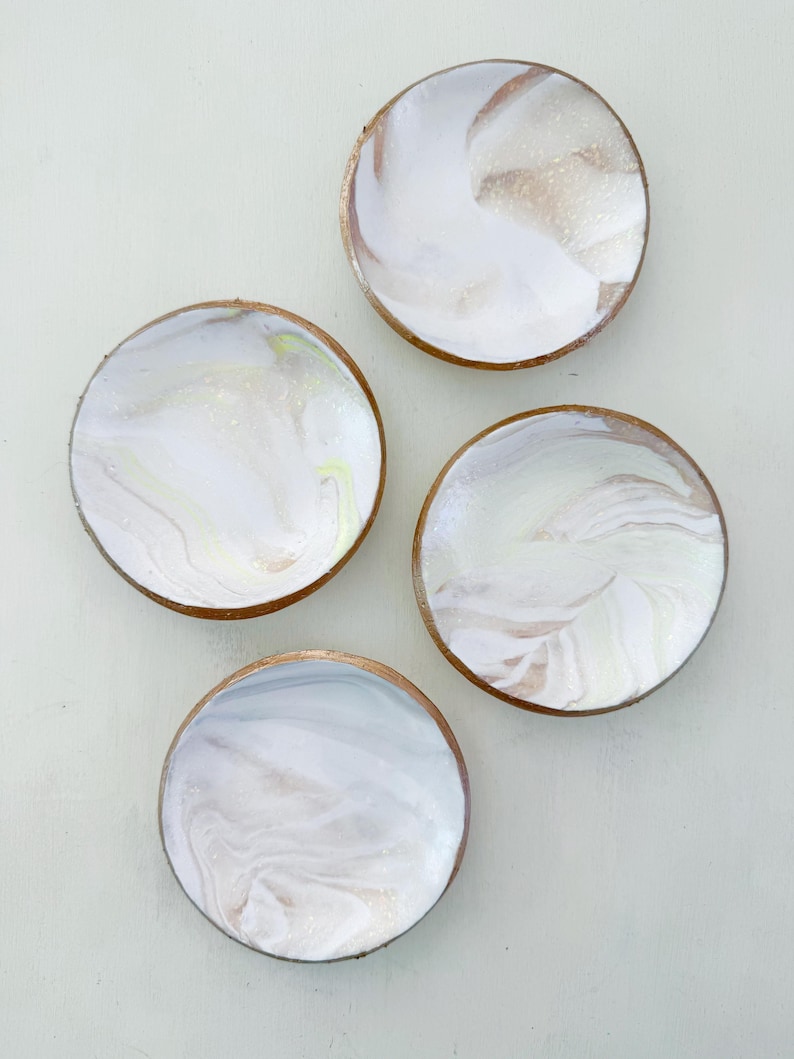 May include: Four round coasters with a white and gold marbled design. Each coaster has a gold-colored rim. The coasters are arranged on a light-colored surface.