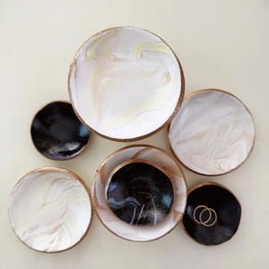 May include: A collection of small, round decorative dishes. The dishes feature a marbled design in white, black, and gold. Some dishes have gold rims. Two gold rings rest on one of the black dishes.