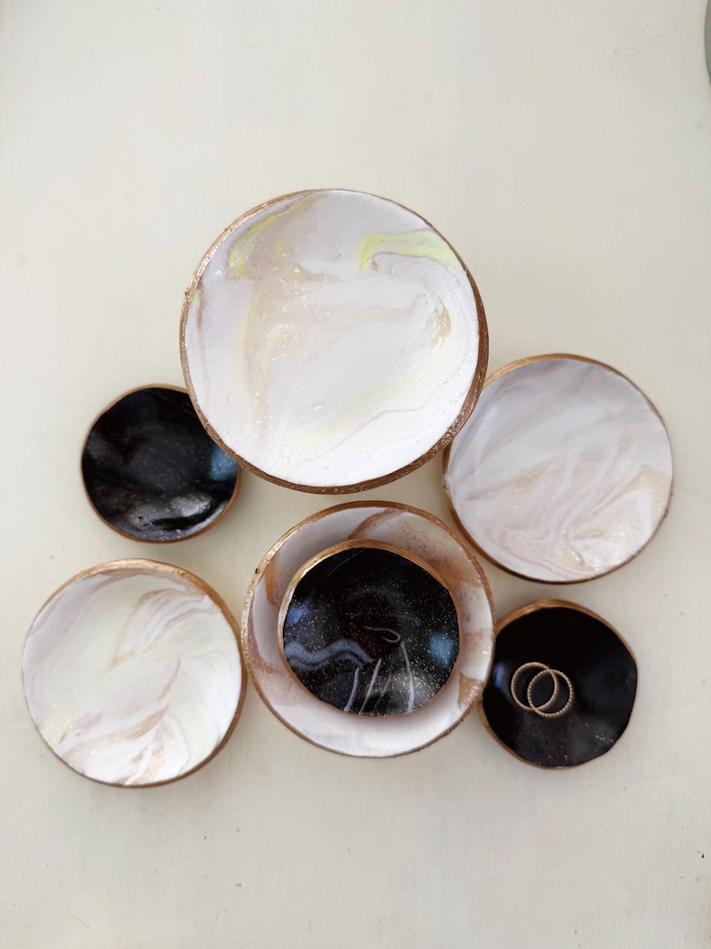 May include: A collection of small, round decorative dishes. The dishes feature a marbled design in white, black, and gold. Some dishes have gold rims. Two gold rings rest on one of the black dishes.