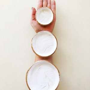May include: Three small, round, white bowls with gold rims are stacked on top of each other. The bowls are filled with white paint and are held by a person's hand and arm. The bowls are of varying sizes.