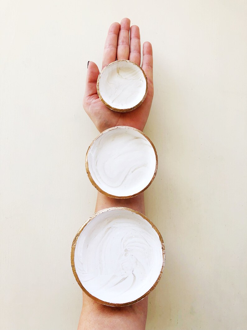 May include: Three small, round, white bowls with gold rims are stacked on top of each other. The bowls are filled with white paint and are held by a person's hand and arm. The bowls are of varying sizes.