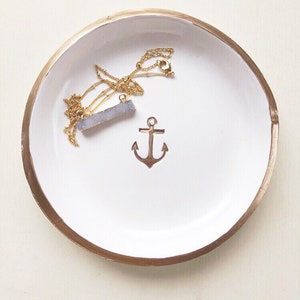 May include: A white ceramic trinket dish with a gold rim and a gold anchor design. A gold necklace with a blue-grey crystal pendant rests inside the dish.