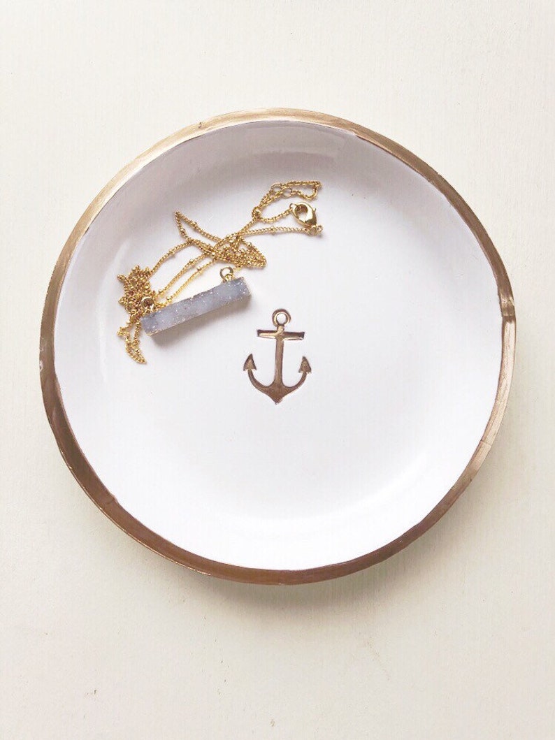 May include: A white ceramic trinket dish with a gold rim and a gold anchor design. A gold necklace with a blue-grey crystal pendant rests inside the dish.