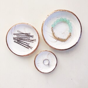 May include: Three decorative white and gold-rimmed trinket dishes with marble-like patterns. One dish holds bobby pins, another a beaded bracelet with a gold-colored fishbone charm, and the smallest dish contains a silver ring.