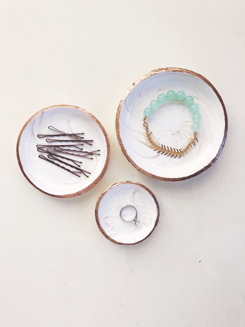 May include: Three decorative white and gold-rimmed trinket dishes with marble-like patterns. One dish holds bobby pins, another a beaded bracelet with a gold-colored fishbone charm, and the smallest dish contains a silver ring.