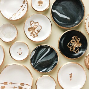 May include: A collection of decorative bowls with gold rims and various designs. Some bowls are white with gold arrows, a seashell, an anchor, or a seahorse. Others feature black and white marbled patterns or a black base with a gold octopus.
