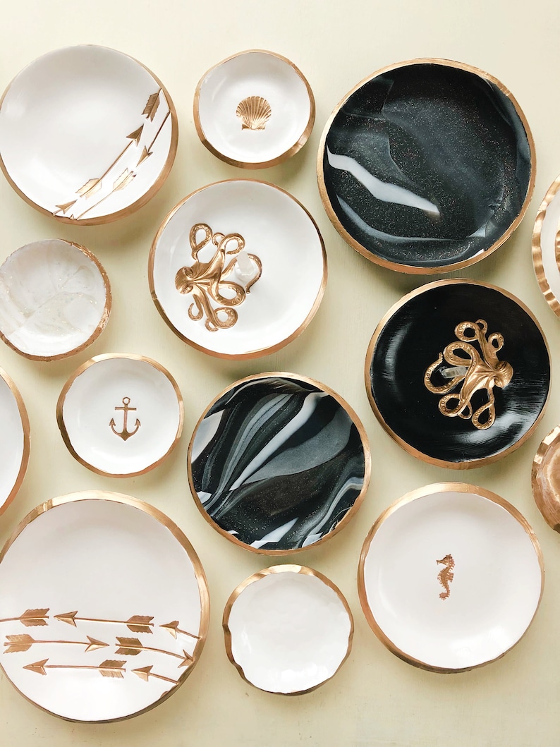 May include: A collection of decorative bowls with gold rims and various designs. Some bowls are white with gold arrows, a seashell, an anchor, or a seahorse. Others feature black and white marbled patterns or a black base with a gold octopus.