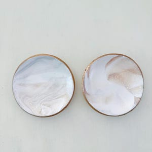 May include: Two round coasters with a marbled design. The coasters have a light blue, white, and gold color scheme. The edges of the coasters are a dark brown color.