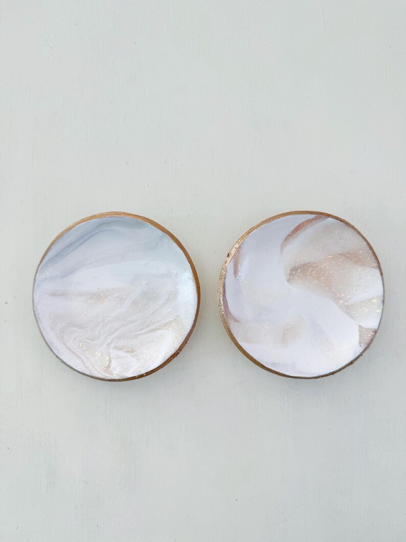 May include: Two round coasters with a marbled design. The coasters have a light blue, white, and gold color scheme. The edges of the coasters are a dark brown color.