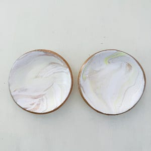 May include: Two round coasters with a white and marbled design. The coasters have a light brown wooden edge. One coaster has a swirl of white, tan, and pink. The other coaster has a swirl of white, yellow, and tan.