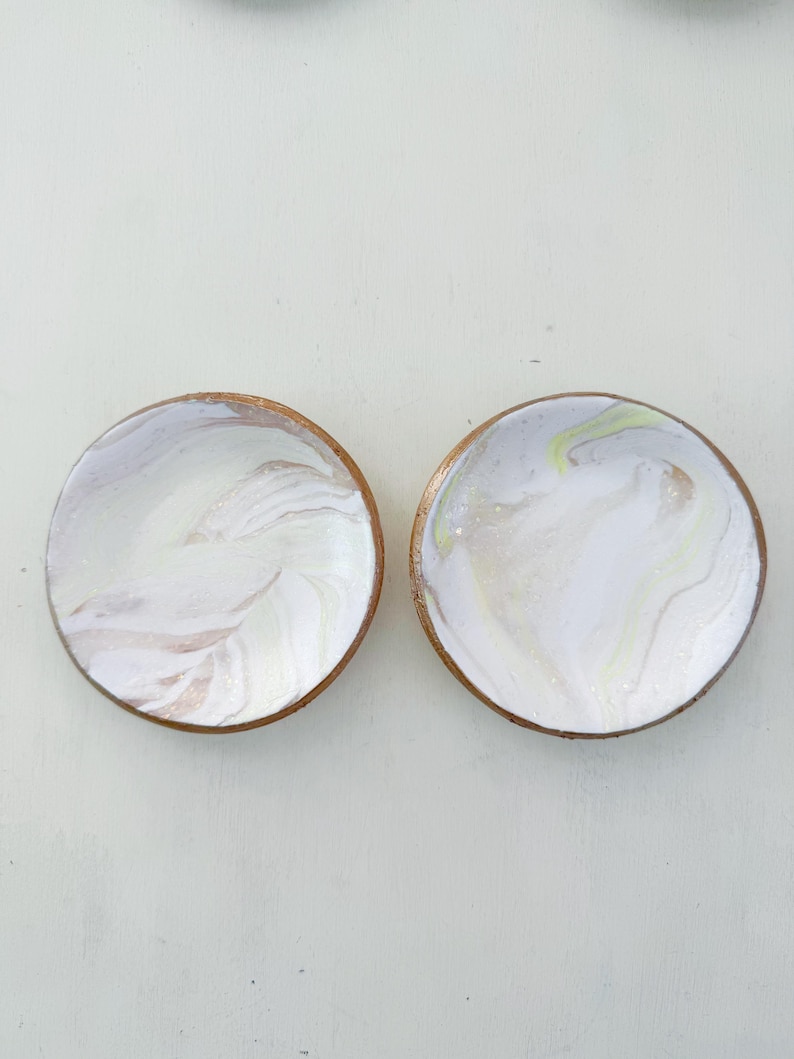 May include: Two round coasters with a white and marbled design. The coasters have a light brown wooden edge. One coaster has a swirl of white, tan, and pink. The other coaster has a swirl of white, yellow, and tan.