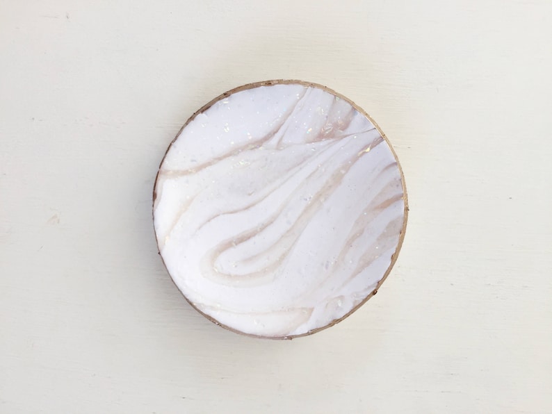 May include: A round, decorative bowl with a white and beige marbled design. The bowl has a natural, light brown rim. The interior features a swirling pattern of white and beige, creating a unique, artistic look. This bowl is perfect for holding small items.
