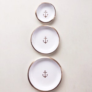May include: Three white ceramic dishes with a gold rim and a brown anchor design in the center. The dishes are arranged in a vertical line, decreasing in size from bottom to top. Nautical themed decor.