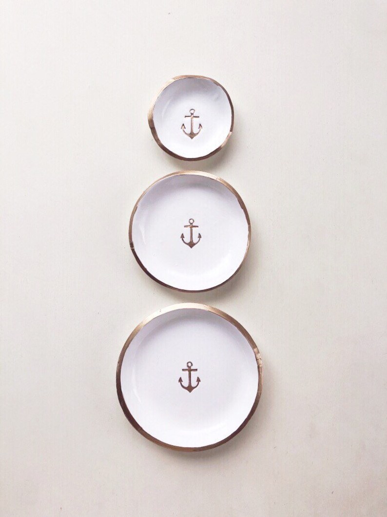 May include: Three white ceramic dishes with a gold rim and a brown anchor design in the center. The dishes are arranged in a vertical line, decreasing in size from bottom to top. Nautical themed decor.