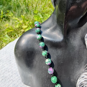 May include: A necklace featuring green and pink gemstone beads. The beads are round and strung together with black string. The necklace is displayed on a black mannequin bust against a backdrop of green foliage.