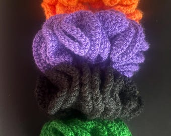 Può includere: Un set di quattro elastici per capelli all'uncinetto impilati verticalmente. Gli elastici sono di colore arancione brillante, viola vivace, nero solido e verde intenso. Ogni pezzo presenta una struttura in filato testurizzato e arricciato.