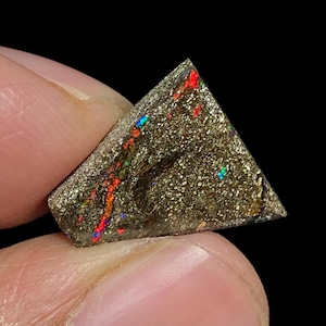 May include: A triangular-shaped gemstone with a dark, speckled base and vibrant flashes of red, orange, and blue. The stone is held between fingers against a black background, highlighting its natural beauty.