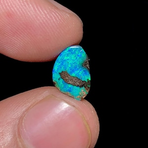 May include: A small, irregular-shaped boulder opal gemstone featuring vibrant flashes of electric blue and green color play across its surface, contrasted by patches of dark, earthy brown matrix material throughout the stone's textured face.