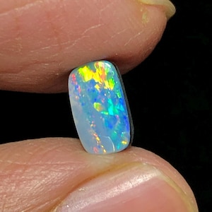 Könnte beinhalten: Ein rechteckiger Opal-Edelstein mit hellblauer Grundfarbe. Der Opal zeigt lebhafte Blitze in Gelb, Orange, Grün und Blau, wodurch ein farbenfroher, schillernder Effekt entsteht. Der Stein wird zwischen zwei Fingern vor schwarzem Hintergrund gehalten.
