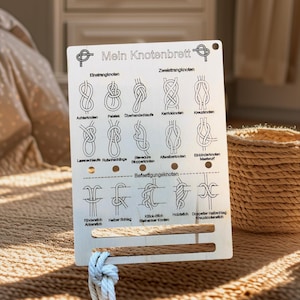 Op de afbeelding: Houten knoopbord met diverse knoopdiagrammen en de Duitse tekst "Mein Knotenbrett". Het bord toont diagrammen van verschillende knopen, waaronder de Elnetrangknotan en de Zweletrangknotan. Een touw is aan de onderkant van het bord bevestigd.