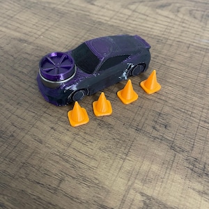 May include: A 3D-printed toy car in black and purple with a prominent purple circular wheel-like component on the hood. The car is accompanied by four small, bright orange traffic cones arranged in a line next to the vehicle body.