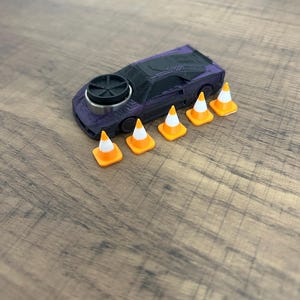 May include: A purple and black toy sports car featuring a circular black component on the hood, accompanied by five miniature orange and white traffic cones arranged in a row alongside the vehicle on a wooden surface.