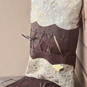 May include: Cushion with a cowhide pattern in white and gold, featuring a dark brown leather corset design. The pillow has a fringe trim at the bottom and is propped against a wooden surface.