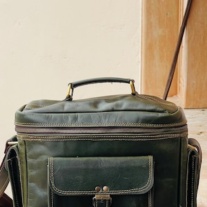 May include: A dark olive green leather camera bag featuring a top handle, a main zippered compartment, and a front pocket with a buckle closure. The bag includes side pockets and is accented with visible contrast stitching and brass hardware.