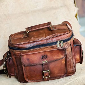 Puede incluir: Un bolso de cuero marrón con múltiples compartimentos, un asa superior y una correa para el hombro. El bolso presenta una solapa frontal con cierre de hebilla y las iniciales "JG" en relieve en la parte delantera. El bolso tiene cierre de cremallera y bolsillos laterales.