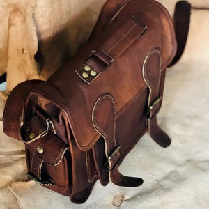 Puede incluir: Una bandolera de cuero marrón con múltiples compartimentos y hebillas de color latón. La bolsa tiene un cierre de solapa y correas ajustables. El cuero tiene un aspecto envejecido, lo que le da un aspecto vintage. La bolsa está diseñada para transportar objetos.