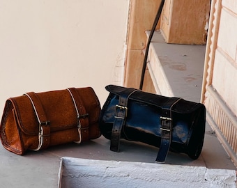 May include: Two leather bags, one brown and one black, are displayed. The brown bag has a textured appearance with white stitching and brass buckles. The black bag has a glossy finish and similar brass hardware. Both bags are rectangular with a curved top.