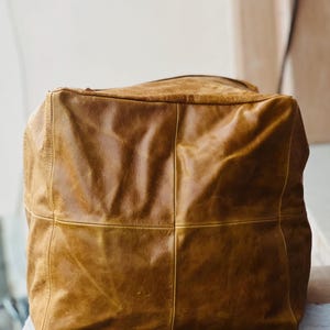 May include: A large, square-shaped, brown leather bag. The bag is constructed from several panels of leather, with visible stitching. The leather has a distressed appearance, with variations in colour and texture, giving it a vintage look. The bag appears to be empty.