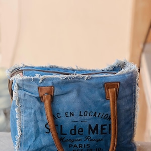 May include: A blue tote bag with brown leather handles and trim. The bag has a distressed, frayed edge design and features black text that reads "SEL de MER" and "PARIS".