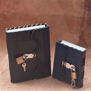 May include: Two black journals with textured covers, scalloped edges, and white pages. Each journal features a metal latch secured by a small brass padlock with a silver key hanging from it. The larger journal has a striped pattern on the spine.