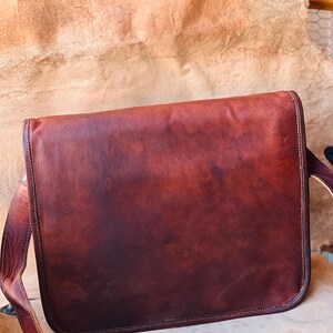Puede incluir: Un bolso mensajero de cuero marrón con cierre de solapa y correa ajustable para el hombro. El bolso tiene un color marrón rojizo intenso con detalles visibles de grano y costura. El bolso está diseñado para transportar objetos.