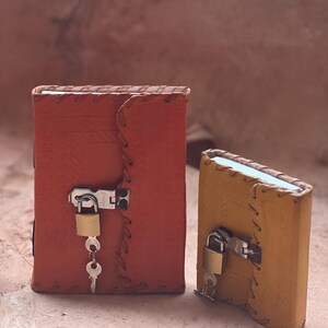 May include: Two rectangular leather-bound journals, one larger orange and one smaller yellow, each featuring embossed patterns, stitched edges, and a metal latch secured with a small brass padlock and a single silver key hanging from the lock.