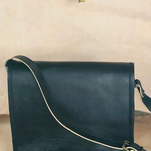 Puede incluir: Un bolso mensajero de cuero verde oscuro con cierre de solapa y correa de hombro ajustable. La correa tiene una franja de acento color crema. La bolsa presenta una hebilla y herrajes metálicos. La bolsa está diseñada para transportar objetos.