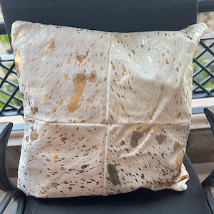 Könnte beinhalten: Quadratisches Zierkissen mit weißem und goldenem Kuhfellmuster. Das Kissen hat ein Patchwork-Design mit goldenen Metallic-Akzenten. Das Kissen ist quadratisch und scheint aus einem weichen Material zu bestehen.