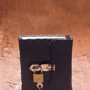 May include: A dark blue leather journal with an embossed pattern and a stitched edge. The journal features a silver-toned metal latch secured by a small brass padlock with a set of keys hanging from the bottom, keeping the white pages enclosed.