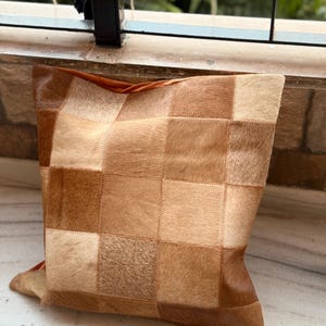 May include: A square cushion with a patchwork design. The cushion features a variety of brown and tan squares, creating a textured appearance. The cushion is made of a soft material and is suitable for home decor.