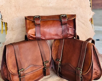 May include: Three brown leather bags with brass buckles. The bags vary in shape and size, with two having a saddlebag design and one rectangular. The leather has a rich, warm tone, and the bags appear to be handmade.