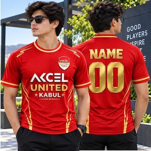 May include: A red sports jersey with gold trim, featuring the text AKCEL UNITED KABUL on the front with a team crest. The back displays the word NAME and the number 00 in gold lettering. The shirt has short sleeves and a round neckline.