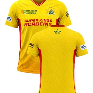May include: A yellow V-neck jersey with red side panels and trim. The front features the text North Texas Food Bank, Texas Super Kings, and Super Kings Academy. The back displays the hashtag Whistle Podu. Logos for ODC and DTA are visible.