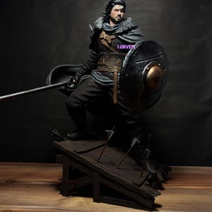 May include: A detailed action figure of a warrior with dark hair and a beard, wearing a grey fur-collared cloak, a brown leather corset, and dark trousers. The figure holds a sword and a round black shield, standing on a wooden platform with arrows.