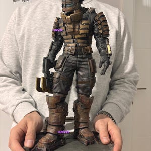 May include: A detailed resin figure of a space suit, featuring a helmet and armor in shades of brown, black, and bronze. The figure is holding a weapon and is standing on a rectangular base. The text "High quality Resin figure" is visible.