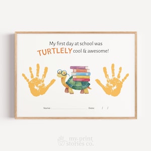 May include: A framed print with the text "My first day at school was TURTLELY cool & awesome!". It features a cartoon turtle wearing glasses and carrying books, with orange handprints on either side. There are spaces for a name and date.