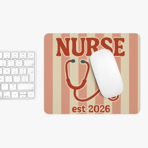 May include: A rectangular mousepad with a retro design featuring the word "NURSE" in brown letters, a red stethoscope graphic, and the text "est 2026". A white wireless mouse rests on the pad, with a white keyboard in the background.