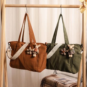 Könnte beinhalten: Zwei Cord-Tote-Bags, einer in Rost und der andere in Olivgrün, hängen an einem Holzgestell. Jede Tasche hat einen dekorativen Riemen und ein kleines Stofftier. Die Taschen haben eine Tasche auf der Vorderseite.