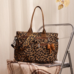 Può includere: Una borsa tote con stampa leopardata dotata di due manici in tessuto marrone, una tracolla regolabile in tinta, diverse tasche esterne con cerniera e un piccolo ciondolo a forma di fiocco marrone fissato a uno dei cursori metallici argentati.