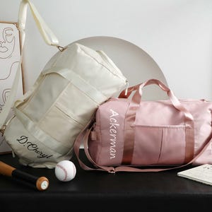 Könnte beinhalten: Zwei personalisierte Sporttaschen, eine cremefarbene und eine rosa, mit Schultergurten. Die cremefarbene Tasche hat "D. Cheryl" in Schrift, und die rosa Tasche hat "Ackerman". Ein Baseballschläger und ein Ball sind im Vordergrund.